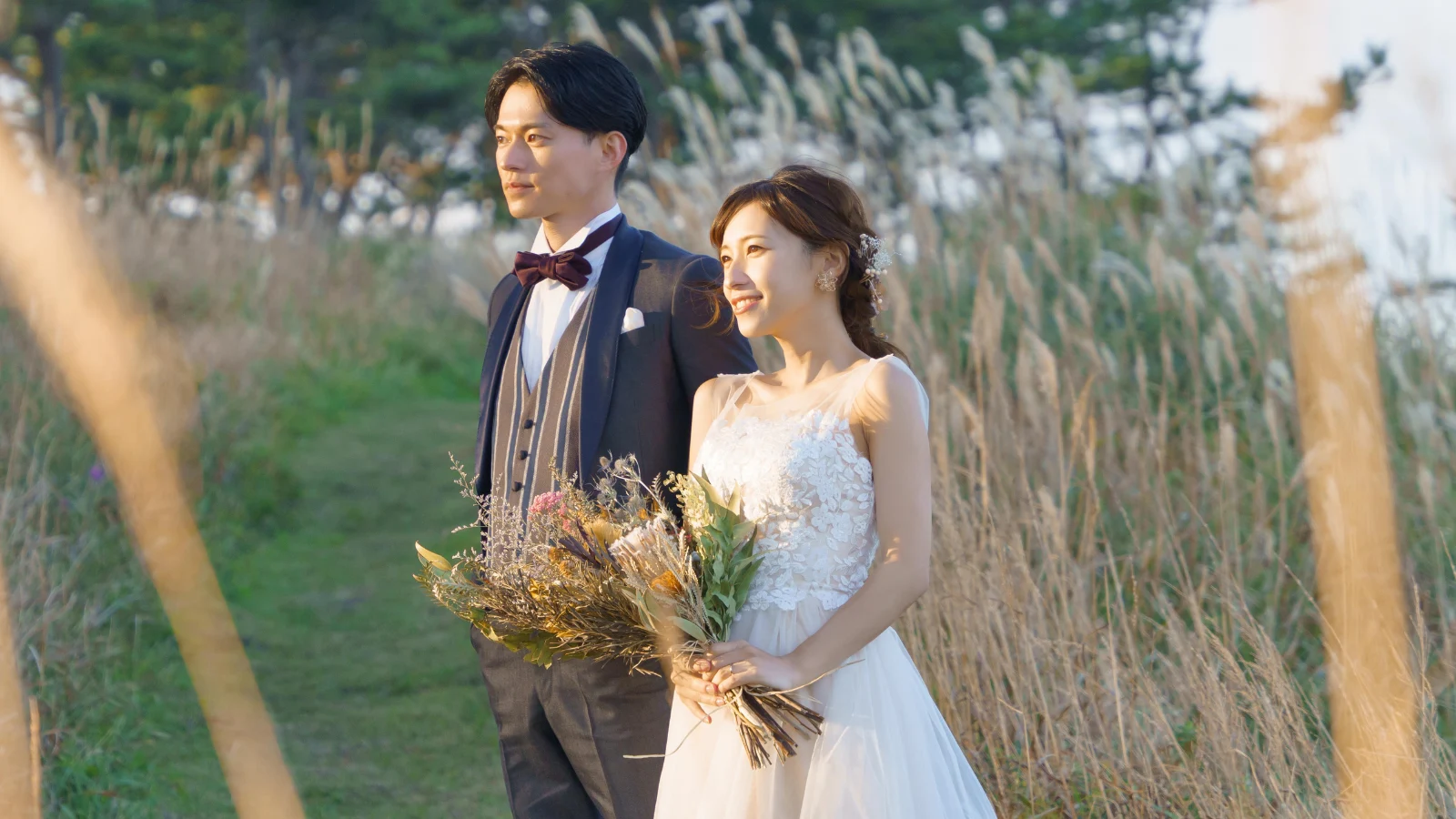 緑とススキに囲まれ、花束を持つドレス姿の花嫁とタキシード姿の新郎