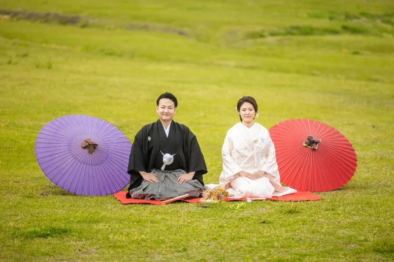 緑の芝生で和傘とともに座る白無垢と紋付姿のおふたり