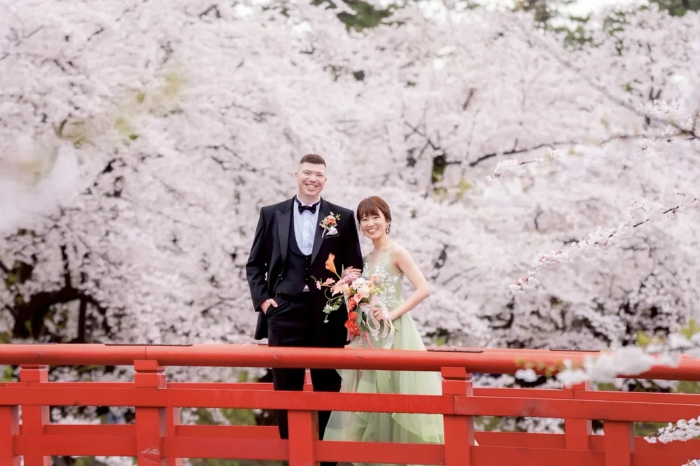 桜の弘前公園の赤い橋で並ぶウェディングドレスとタキシード姿の新郎新婦